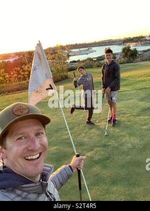 Vacation in New Port Beach on golf course with dad, son and cousin, holding a flag on the green with a lake in background during sunset Stock Photo