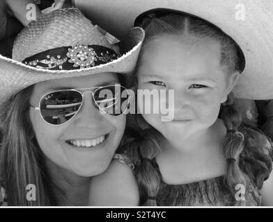 Woman wearing aviator sunglasses and young girl, both wearing cowboy hats while at the rodeo smiling. Black and white photo.