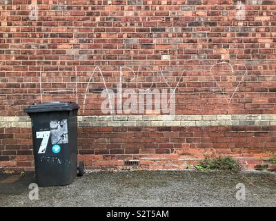 A black wheelie bin stands in front of the brick wall of a house with the word happy and a heart drawn on the wall in white chalk Stock Photo