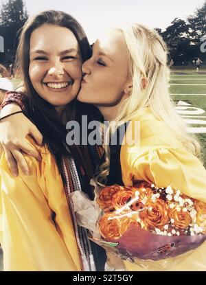 Graduation Day for two beautiful girls. They are wearing yellow gowns and hugging and giving a kiss on the cheek. One is holding flowers Stock Photo