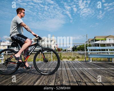 Man with tattoos Cycling on boardwalk with beach houses , Ocean City, New Jersey, USA Stock Photo