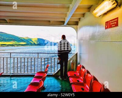 Man standing alone on deck of ferry near rows of empty red plastic seats, Inner Hebrides, Scotland Stock Photo