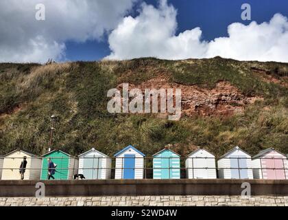 Row of colourful beach huts with two people walking their dog along promenade, Seaton, Devon, UK Stock Photo