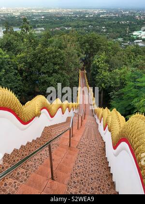 Steps with the Golden Naka statue on each side of the stairs leading down through a forest at Wat Phra That Doi Kham in Chiang Mai. Stock Photo