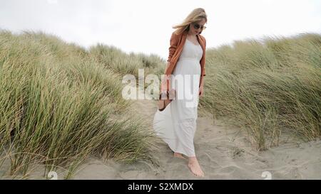 Young blonde woman in long white dress walking down sandy hill