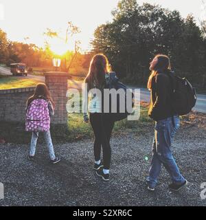 Three school age girls waiting for school bus as it approaches in the early morning at sunrise Stock Photo