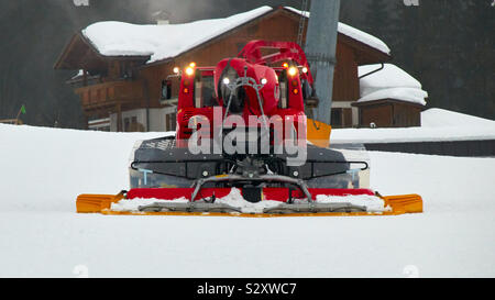 A red snow groomer starts to prepare the ski slopes for its mission. The machine from Tyrol in Austria can be seen from the front and has its lights on. Stock Photo