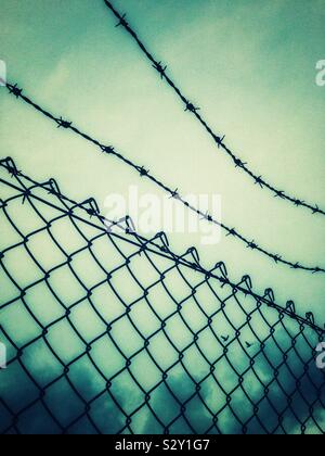 Mesh fence and barbed wire against cloudy sky. Stock Photo