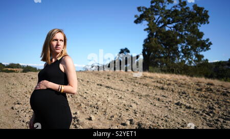 Young blonde pregnant woman wearing black looking over her shoulder with serious expression on dirt covered hill with large fallen tree in background Stock Photo