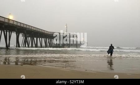 PISMO BEACH, CA, OCT 2019: surfer in black wetsuit walks out of Pacific Ocean next to the pier, on a foggy late afternoon Stock Photo