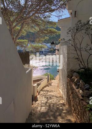 The coastal footpath goes between whitewashed houses down to Sa Tuna beach basking in November sunshine near Begur, Costa Brava in Spain. Stock Photo