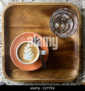 Cup of espresso coffee with attractive pattern on froth. Served on orange saucer, on small wooden tray with a glass of water Stock Photo