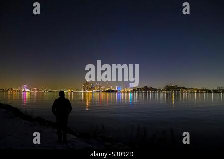 Silhouette of young adult man during a winter night viewing a river and city night life. Detroit, Ambassador Bridge, and Canada in view. Stock Photo