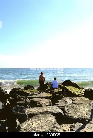 Two boys sitting on rocks contemplating the ocean. Stock Photo
