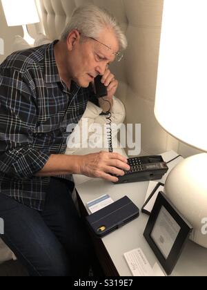 Man on a hotel room telephone making a call to the front desk with his cellular iphone on the table. Stock Photo