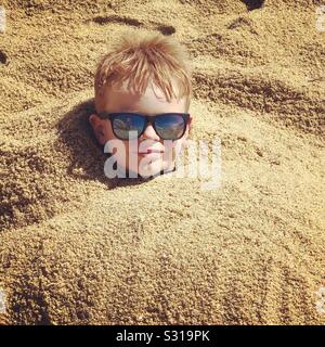 Boy wearing sunglasses relaxing on beach whilst buried in sand Stock Photo
