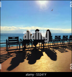 A man and 2 women sit on chairs in the bright sunshine watching the sun start to set over the Mediterranean Sea. Interesting shadows and a well placed seagull! Photo ? COLIN HOSKINS. Stock Photo