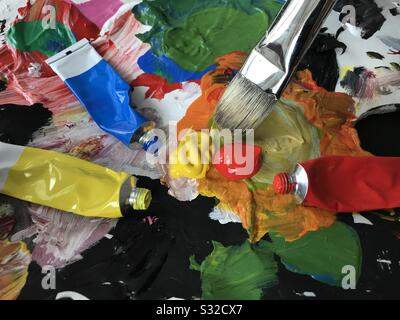 Artist palette with paint brush mixing red and yellow colors and paint tubes scattered around. Stock Photo
