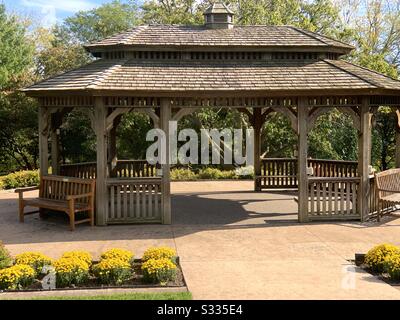 DUBUQUE, IOWA, Fall 2019--Landscape photo of rustic wooden gazebo on patio under a blue sky surrounded by trees and bright yellow mums on a fall day in an arboretum. Stock Photo