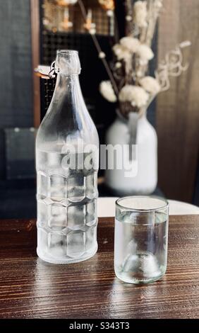 Simple glass bottle of water alongside a glass of water on a table with a vase of flowers in the background. Stock Photo