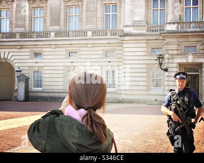 A young woman is seen from behind, tourist taking photographs at the gates to Buckingham Palace, London as an armed police officer watches on with an assault rifle Stock Photo