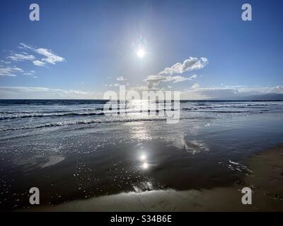 Centered landscape view of low sun over the Pacific Ocean with sparse clouds in blue sky, gentle waves lapping sandy beach in foreground Stock Photo