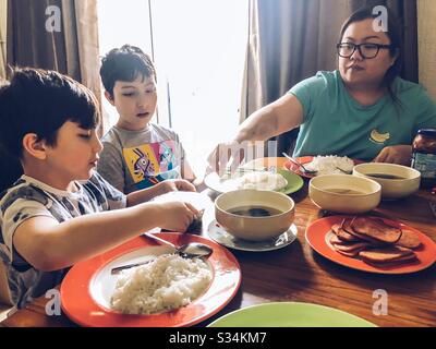 A family eating a meal together at a dining table. The table is covered with bowls and plates with rice, meat and soup. Stock Photo