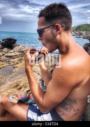 A young man relaxing by the sea drinking Maté on the rocky shoreline. Stock Photo