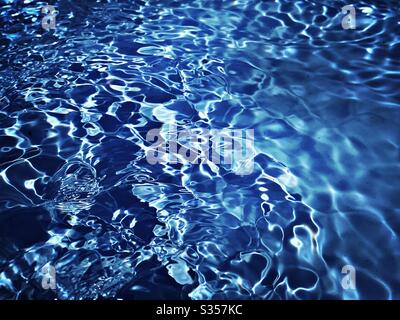 A close up view on the surface of water in a blue swimming pool. Light reflects off gentle ripples creating a colourful summer background pattern. Fresh, clean and vibrant summery vibes. Stock Photo