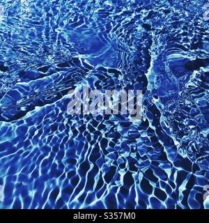 A close up view on the surface of water in a blue swimming pool. Light reflects off gentle ripples creating a colourful summer background pattern. Fresh, clean and vibrant summery vibes. Stock Photo
