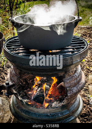 Cast iron cooking pot bubbling on top of wood fires outdoor stove made from old car wheels Stock Photo