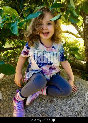 Happy little girl sitting on a rock under an Apple tree. Stock Photo