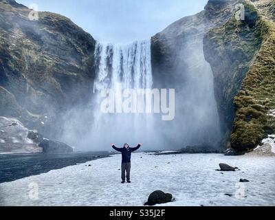 Man standing in front of a majestic waterfall in Iceland with his arms in the air. Snow covering the ground. Stock Photo
