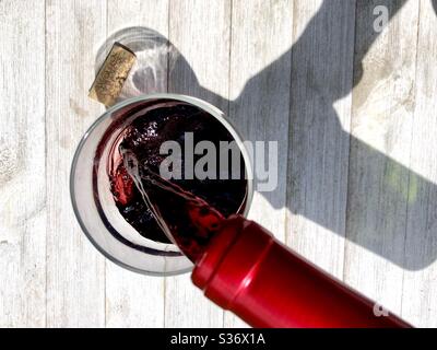 Red wine being poured into a glass on a wooden surface in natural sunlight Stock Photo
