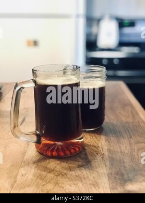 Two glasses of home made dark beer sit on a wood counter in a kitchen.