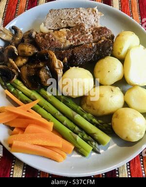 Steak dinner with new potatoes carrots asparagus and mushrooms served on a pale grey plate with a vibrant, orange background Stock Photo