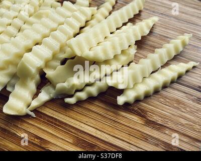 Preparing crinkle cut fries (US) or chips (UK) in the kitchen on a rustic wooden chopping board. Sliced potatoes with jagged edges ready for frying Stock Photo