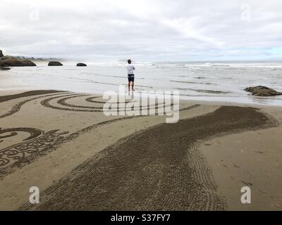 A man looking out at the ocean while the rising tide erases a sand labyrinth, in Bandon, Oregon. Stock Photo