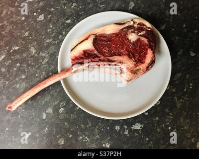 Raw red Tomahawk steak on a duck egg blue plate with a black worktop background.  Copy space on the plate for text Stock Photo