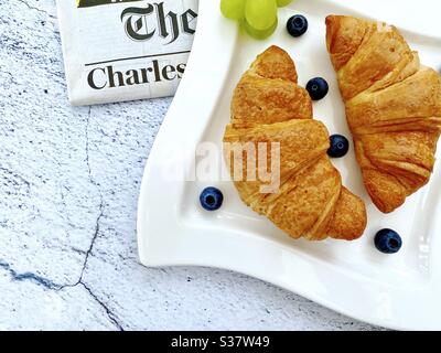 Closeup view of freshly baked croissants on a white plate with blueberries and grapes. Tasty breakfast with morning newspaper on a natural granite stone table. Stock Photo
