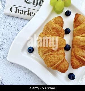 Closeup view of freshly baked croissants on a white plate with blueberries and grapes. Tasty breakfast with morning newspaper on a natural granite stone table. Stock Photo