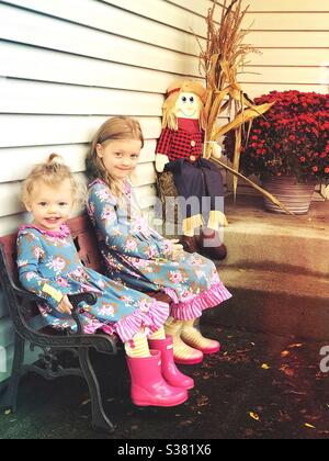 Sisters wearing matching outfits sitting on a wooden kids bench next to a scarecrow sitting on hay in autumn Stock Photo