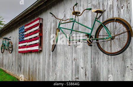 WATERVILLE, IOWA, July 15, 2020–Closeup photo of wooden brightly painted American flag between two green rusted bicycles built for two hanging on the side of a gray washed out wooden building. Stock Photo