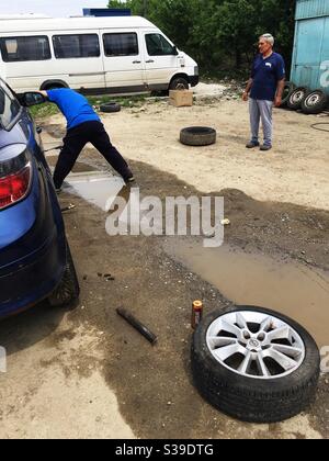 Changing tires on a car at a service shop in the Romanian countryside, with muddy water, detached wheel and bystander in the frame Stock Photo