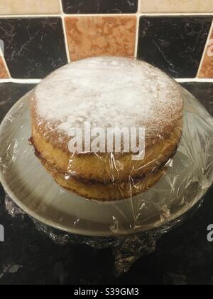 Home baked Victoria Sandwich sponge cake on a plate and covered with cling film, plastic food wrap Stock Photo