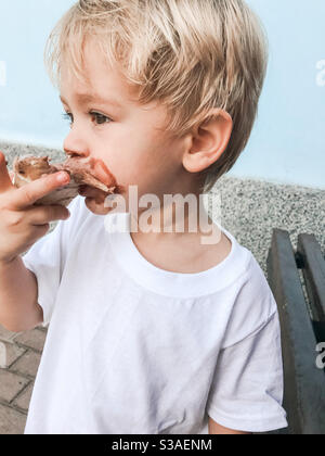 2 year old blonde boy eating chocolate ice cream with a white t shirt on outside on a hot and sunny day Stock Photo