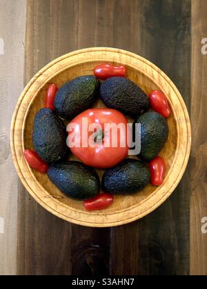 Six Marzano tomatoes, six avocados and one large red tomato on a wooden bowl from above. Stock Photo