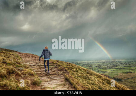 Girl walking up Pendle Hill with rainbow over fields in distance Stock Photo