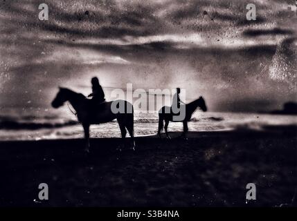 Silhouette of two horse riders on their horse by the sea. Image is edited to give it a vintage tintype photography look. Stock Photo