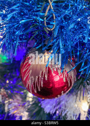 Shiny red Christmas bauble with sparkling gold glitter pattern hanging on a Xmas tree with blue tinsel Stock Photo